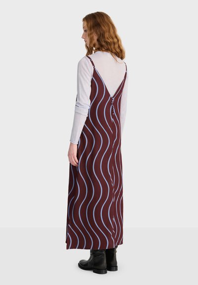 Femme aux cheveux roux ondulés portant une robe marron sans manches avec des rayures ondulées bleues par-dessus une chemise blanche à manches longues et des bottes noires, debout de profil.