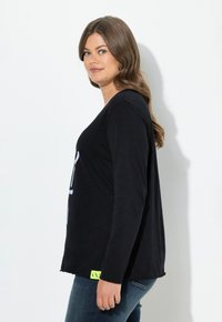 Schwarzes Langarmshirt mit lockerer Passform, aus weichem Stoff, mit sichtbarem Logo am Saum und kombiniert mit blauen Jeans.