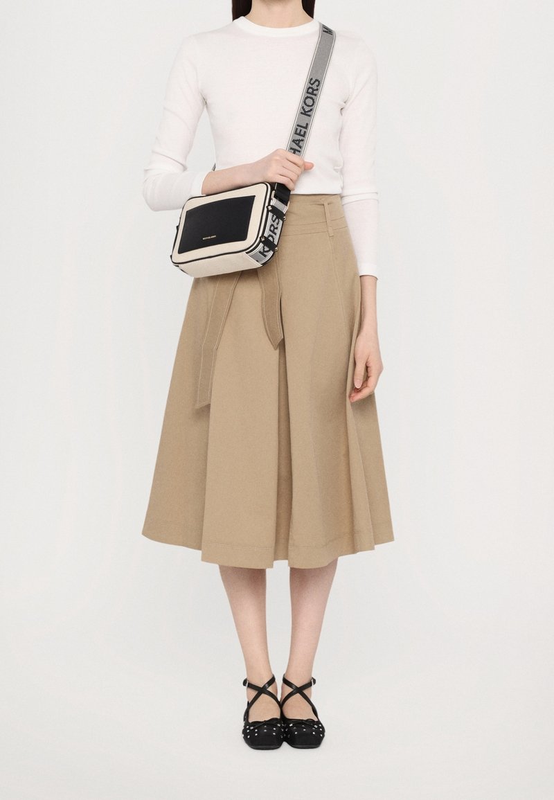 Femme portant un haut blanc à manches longues, une jupe midi beige avec un nœud à l'avant, des ballerines noires à brides croisées cloutées, et un sac à bandoulière beige et noir Michael Kors.