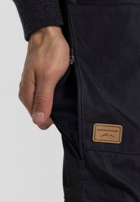 Black pants with a zippered pocket, featuring a tan leather label with "SWEDEMOUNT" and mountain graphic. Textured material, slim design.