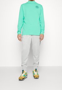 Mint green long-sleeve shirt with a round logo, gray tapered pants with side pockets, and yellow and teal athletic shoes.