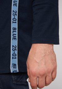 Camicia a maniche lunghe blu navy con una striscia laterale azzurro chiaro che presenta la scritta "25-01 BLUE" in testo blu scuro; vestibilità aderente, materiale misto cotone.