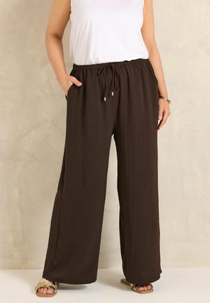 Personne portant un pantalon large marron foncé à taille élastique avec cordon, un haut sans manches blanc et des sandales dorées, debout avec une main dans la poche.