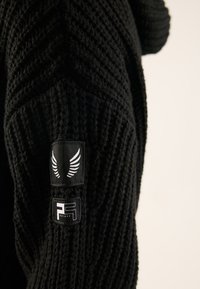 Svart stickad tröja med ett strukturerat mönster, försedda med två logopatchar på ärmen. Materialet verkar tjockt och varmt.