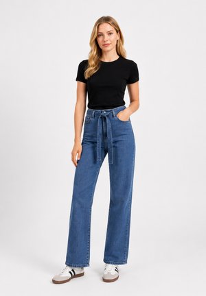 Donna con lunghi capelli biondi che indossa una maglietta nera a maniche corte, jeans blu a vita alta con cintura e scarpe da ginnastica bianche con strisce nere.