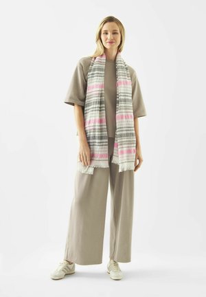 Frau trägt beige weiter Hosen, lockeres beiges Oberteil, weiße Sneakers und einen langen gestreiften Schal mit pinken und grauen Mustern.