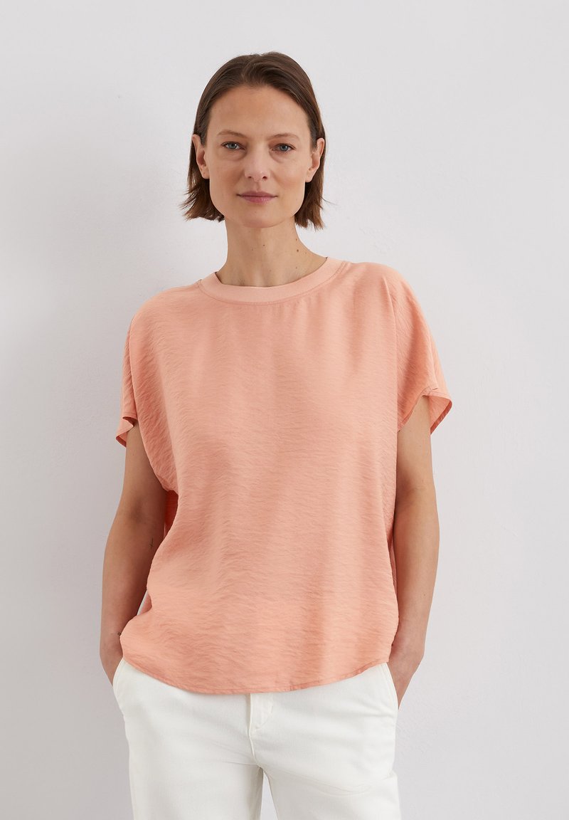 T-shirt à manches courtes de couleur pêche avec une coupe décontractée et un col en rib, présentant un tissu texturé et un design à ourlet arrondi.