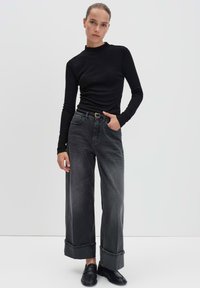 Vrouw in een zwart lange mouwen top, high-waisted wijdvallende opgerolde donkergrijze jeans, zwarte riem en zwarte instappers die tegen een effen achtergrond staat.