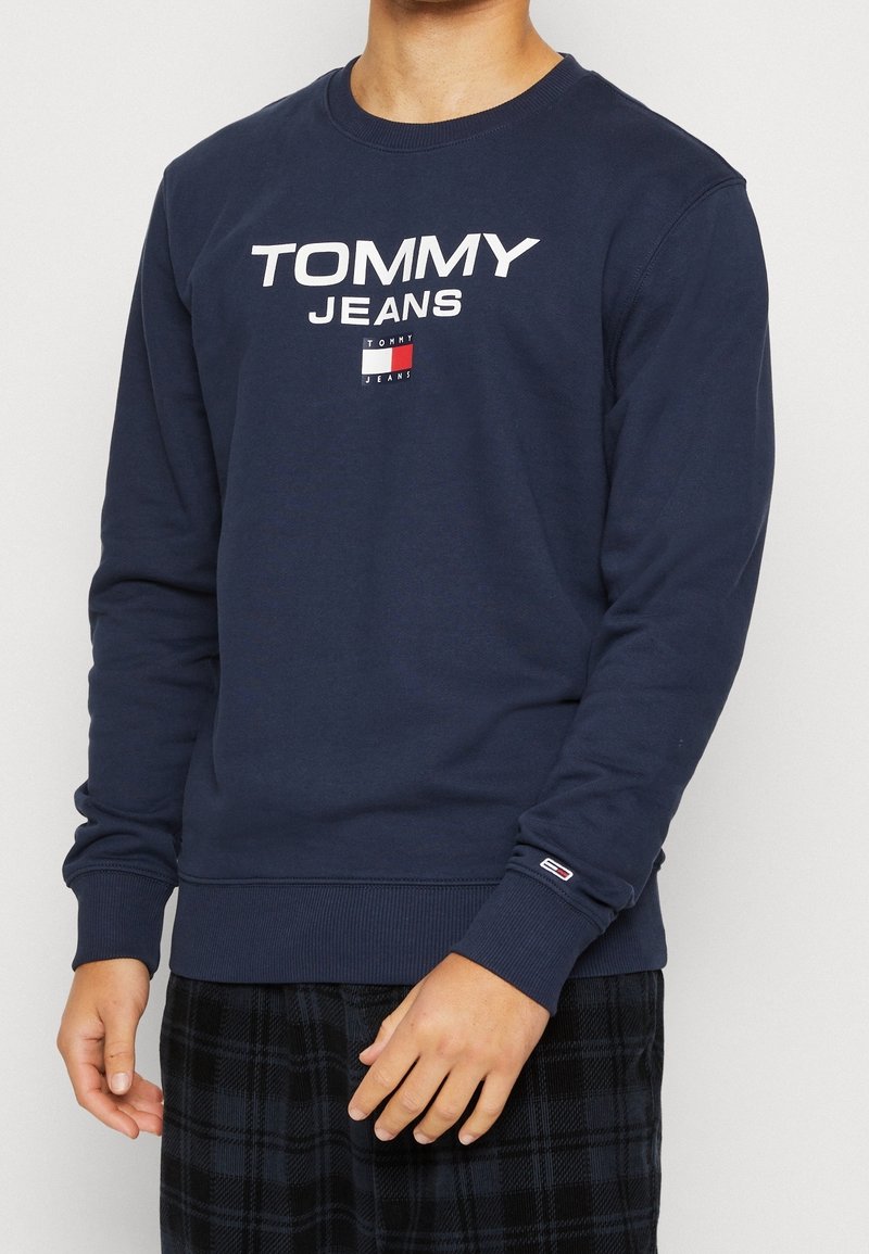 Námořnická modrá mikina s bílým logem "TOMMY JEANS", žebrované manžety a lem, na hrudníku s malým emblémem v červené, bílé a modré barvě.