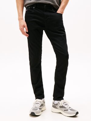 Hombre que lleva pantalones ajustados negros y zapatillas grises y blancas, de pie con una mano en el bolsillo contra un fondo blanco.