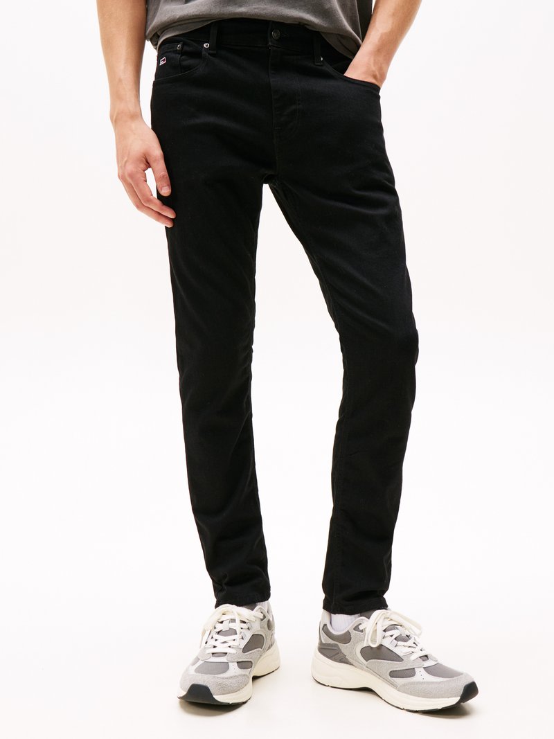 Man in een zwarte slim-fit broek en grijs-witte sneakers, staande met één hand in zijn zak tegen een witte achtergrond.