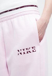 Tuvumā redzams cilvēks, kurš valkā gaiši rozā Nike sporta bikses, ar roku kabatā un redzamu Nike zīmola jostu zem baltas augšdaļas.