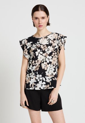 Femme portant un chemisier noir à manches courtes à fleurs beiges et blanches, associé à un short noir, debout devant un fond uni.