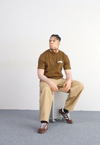 Brun bomulds t-shirt med hvidt logo, khaki cargo bukser og brune sneakers med hvide striber. Sidder på en sølvfarvet metalstol.