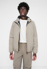 Veste légère beige avec fermeture éclair et capuche, doublure noire, et une bande multicolore le long de la fermeture. Portée par-dessus une chemise blanche.