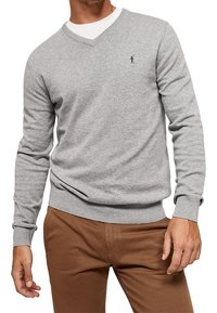 Pull gris à col en V en tissu maille douce, avec manches longues, poignets et ourlet côtelés, et un petit logo sur la poitrine. Associé à un pantalon marron.