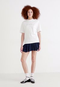 Bílý tričko s nápisem "FEMME", kombinované s námořnickou sukní s volány. Model má na sobě černé baleríny a bílé ponožky. Jednoduchý design a ležérní vzhled.