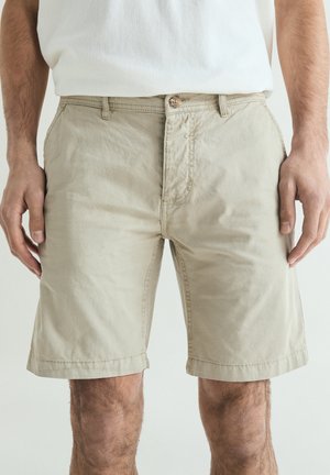 Mand iført beige knælange shorts og hvid skjorte, stående med afslappede hænder langs siden mod en ensfarvet baggrund.
