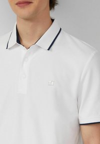 Weiße Poloshirt aus einem strukturierten Stoff, mit einem marineblauen Besatz am Kragen, einer Zwei-Knopf-Leiste und einem kleinen Logo auf der linken Brust.