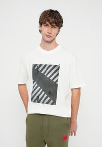 Mladý muž na sebe má biele tričko s čiernobielou grafikou priechodu pre chodcov a olivovo zelené nohavice s červenou záplatou.