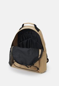 Mochila bege com um forro interior preto, apresentando um compartimento principal e um bolso lateral de malha. Tecido suave, forma estruturada e alças reforçadas.