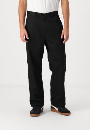 Homme portant un pantalon noir droit, un t-shirt blanc et des baskets noires avec des rayures blanches et des semelles marron, debout devant un fond uni.