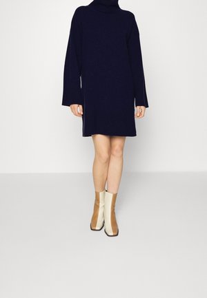 Kvinde iført en mørkeblå, langærmet rullekravesweaterkjole og tofede beige og brune ankelstøvler, stående foran en ensfarvet baggrund.