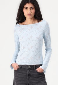 Light blue long-sleeve lace top featuring a floral pattern, boat neckline, and fitted silhouette, paired with dark denim jeans.