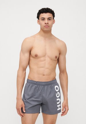 Gray swim shorts with an elastic waistband, featuring a large white "HUGO" logo on the left side. Made of smooth, lightweight material.