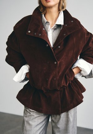 Mujer con camisa blanca de varias capas y traje a cuadros gris bajo una chaqueta de ante marrón oscuro sobredimensionada, con las manos en los bolsillos.