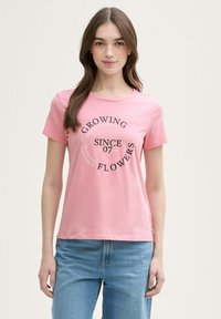 Camiseta de algodón rosa con mangas cortas y cuello redondo. Presenta texto impreso en negro: "CRECIENDO FLORES DESDE 07". Dobladillo recto.