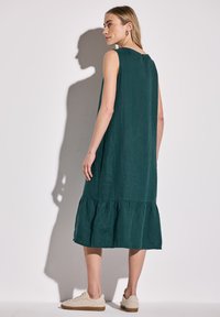 Bezramionowa zielona sukienka lniana z falbaną na dole i zapięciem na guziki z tyłu, teksturowana tkanina, noszona z beżowymi trampkami.