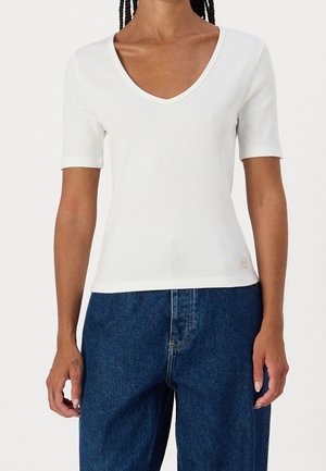 Frau trägt ein eng anliegendes weißes geripptes V-Ausschnitt-Kurzarmshirt und hoch taillierte, locker sitzende dunkelblaue Jeans vor schlichtem Hintergrund.