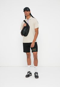 Beige Poloshirt mit lila Reißverschluss, schwarze Shorts mit grüner Schrift, schwarze Kappe und kleiner schwarzer Rucksack; kombiniert mit weißen Socken und schwarzen Schuhen.