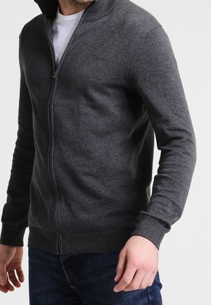 Homme portant un pull zippé gris foncé sur une chemise blanche et un jean bleu devant un fond clair uni.