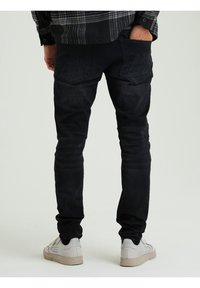 Jeans in denim neri con una vestibilità slim, dotati di tasche posteriori con patte angolate, abbinati a sneakers di colore chiaro. Texture liscia in tutto.