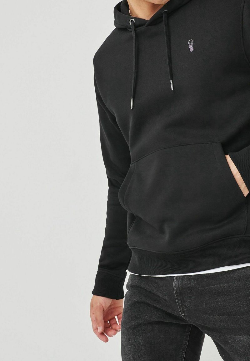 Hombre vestido con una sudadera con capucha negra con un pequeño logo de ciervo, jeans negros y una mano en el bolsillo delantero, contra un fondo liso.