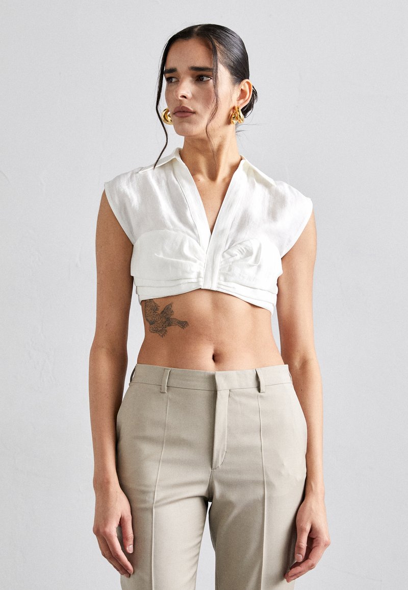 Chemisier blanc court avec un col en V, un devant plissé et des manches courtes, associé à un pantalon beige clair ajusté.