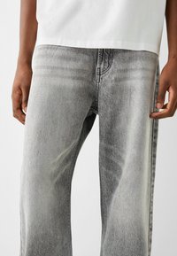 Grå denim jeans med en blekt finish, vid ben-design och synlig sömnad. Tyget har en något grov textur.