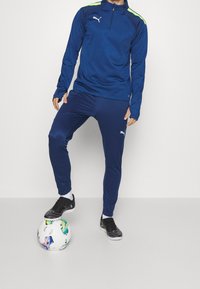 Marineblauw sporttrainingspak met een kwart-rits top, voorzien van neon groene accenten, slim-fit broek en zwarte sportschoenen op een voetbalschoen.