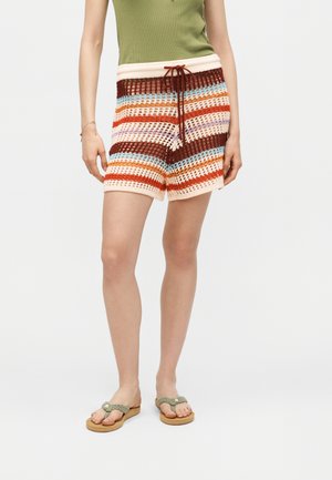 Person iført flerfarvede stribede hæklede shorts, olivengrøn ærmeløs top og beige klipklapper med flettede stropper, stående på hvid baggrund.