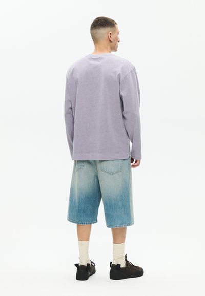 Homme debout, portant une chemise à manches longues violette claire, un short en jean bleu clair, des chaussettes blanches et des baskets noires, vu de dos sur un fond blanc.