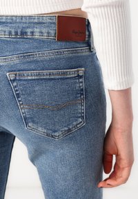 Vaqueros azules de mezclilla con un bolsillo trasero curvado y una parche de cuero marrón con el logo. La tela tiene un acabado texturizado y zonas desgastadas.