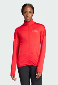 Giacca Terrex rossa con colletto alto, zip frontale completa e maniche lunghe. Tessuto texturizzato e design minimalista, con un logo sul petto.