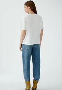 Locker sitzende weiße Bluse mit kurzen Ärmeln, kombiniert mit blauen Gerade-Jeans. Das Model trägt beige Ankle Boots.
