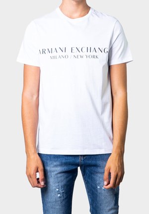 Person trägt ein weißes Armani Exchange T-Shirt mit dem Schriftzug "Milano / New York" und abgenutzte blaue Jeans, dargestellt vom Hals bis zur Hüfte.