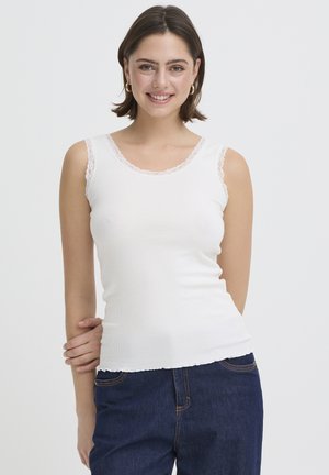 Młoda kobieta w białym bezrękawnym topie z koronkowym wykończeniem i niebieskich dżinsach, uśmiechająca się, pozująca z jedną ręką na biodrze na jednolitym tle.