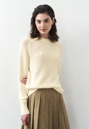 Young woman with dark curly hair wearing a cream knit sweater and olive green pleated skirt, standing against a white background.