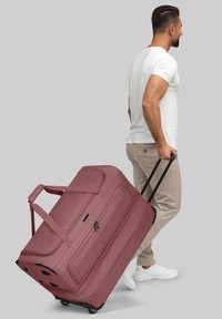 Hombre con camiseta blanca y pantalones beige arrastrando una gran bolsa de viaje de color malva sobre un fondo gris.