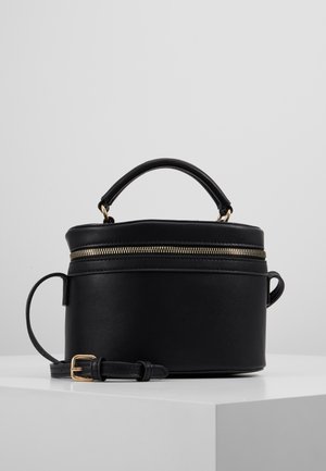 Sac à main ovale en cuir noir avec fermeture éclair dorée, poignée courte et bandoulière réglable posé sur une surface blanche sur fond gris.